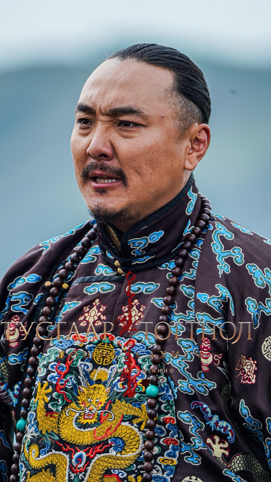 A man in a dark robe embroidered with a golden dragon, prayer beads around his neck