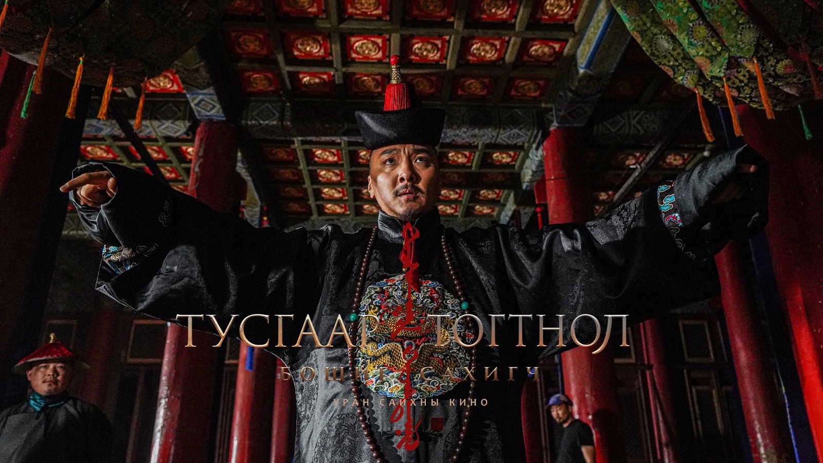 A man in a dark robe with his arms spread wide in a hall of red columns